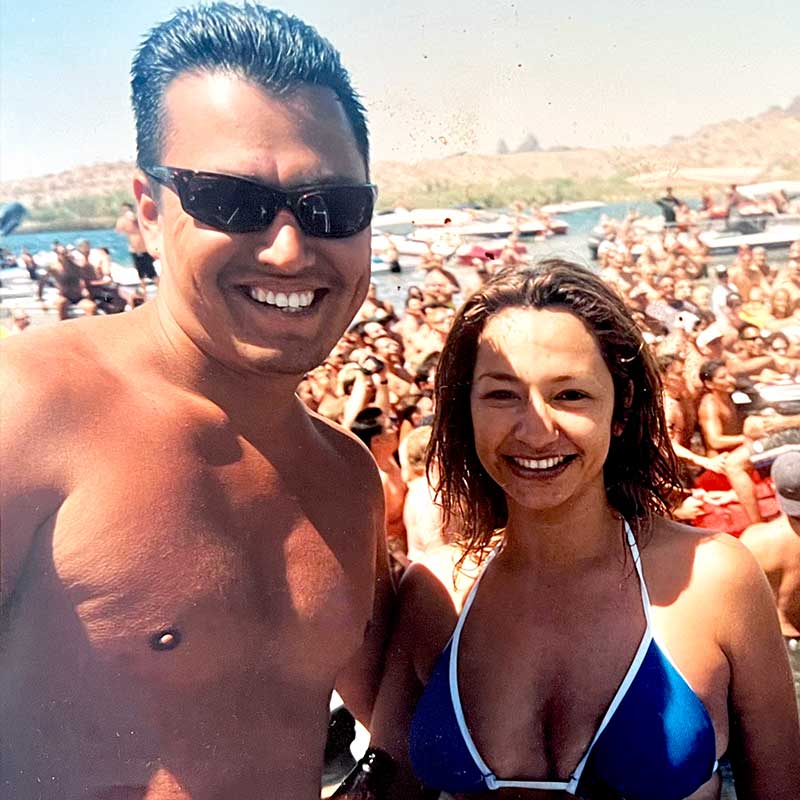 A man and woman in swimwear smile at the camera at a crowded outdoor lakeside event, with boats, people, and a lively Mexican restaurant scene in the background.