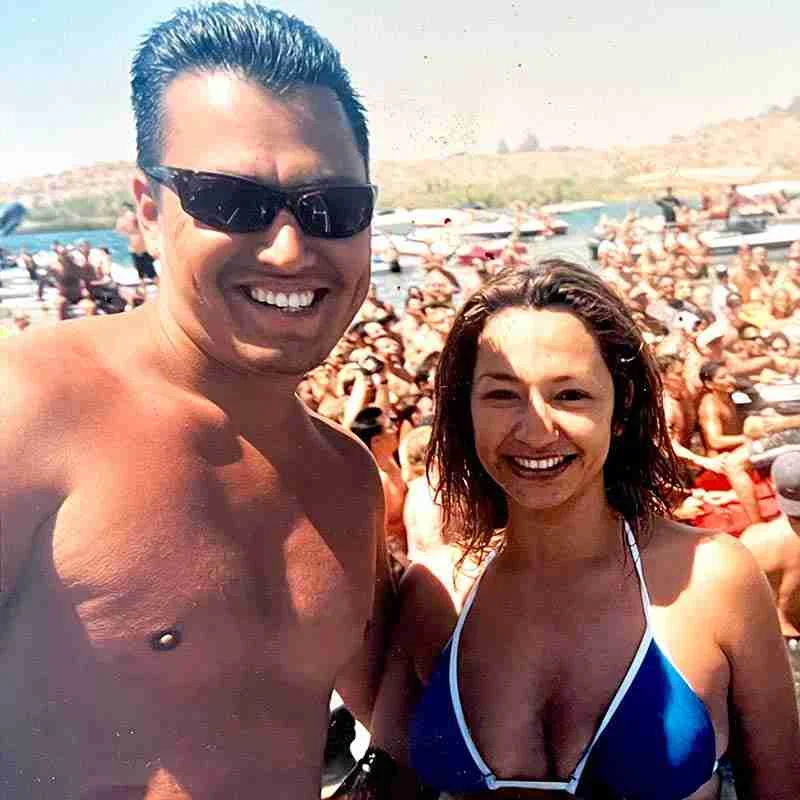 A man and woman in swimwear smile at the camera at a crowded outdoor lakeside event, with boats, people, and a lively Mexican restaurant scene in the background.