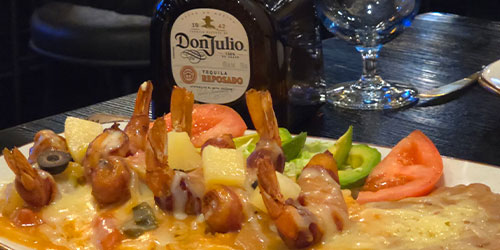 A plate of bacon wrapped shrimp topped with melted cheese, olives, pineapple, and vegetables sits in front of a bottle of Don Julio tequila on a restaurant table.