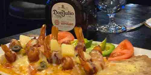 A plate of bacon wrapped shrimp topped with melted cheese, olives, pineapple, and vegetables sits in front of a bottle of Don Julio tequila on a restaurant table.