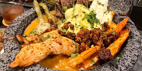 A stone bowl filled with grilled chicken, shrimp, chorizo, avocado, cheese, grilled vegetables, and a red sauce.