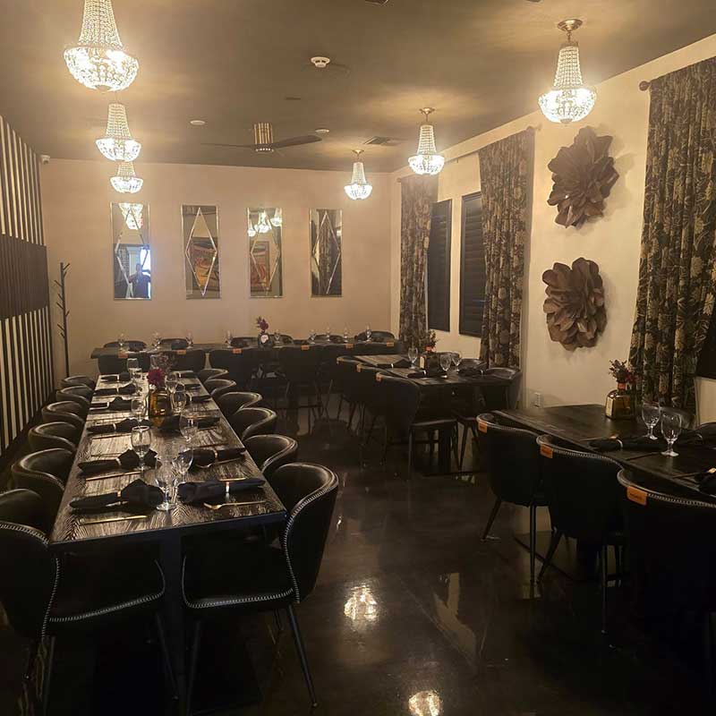 A dimly lit dining room with black tables and chairs, set with plates and glasses, featuring chandeliers, wall art, and curtained windows.