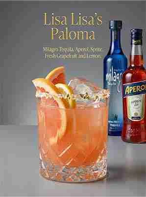 A glass of grapefruit-colored cocktail with ice and fruit garnishes sits in front of Milagro Tequila and Aperol bottles; text lists ingredients for "Lisa Lisa’s Paloma.