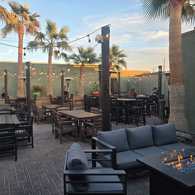 Outdoor patio with palm trees, string lights, wooden tables and chairs, a gray outdoor sofa, and a fire pit, with a few people seated in the background.