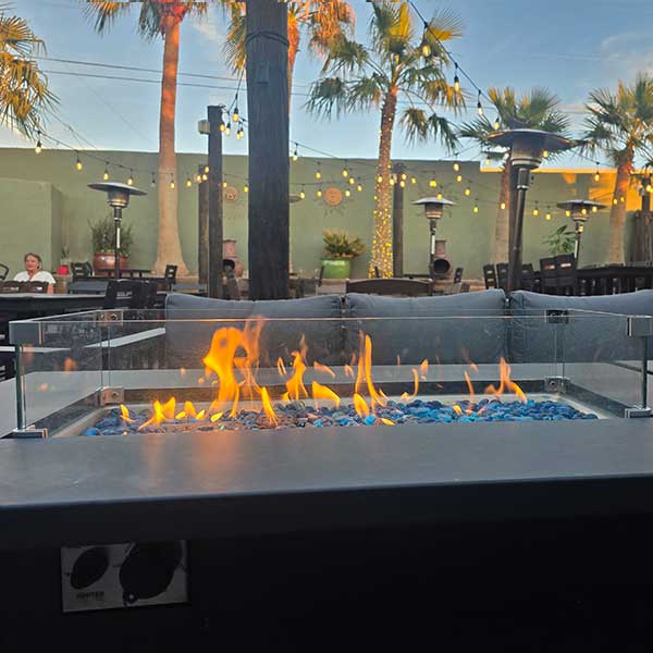 Outdoor patio with a modern fire pit featuring blue glass rocks, surrounded by seating and palm trees, with string lights overhead at dusk.