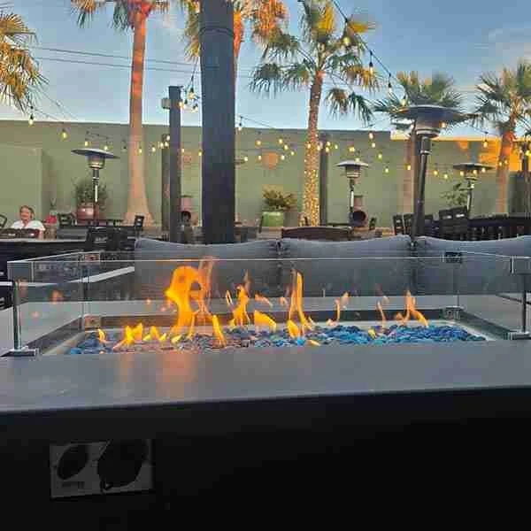 Outdoor patio with a modern fire pit featuring blue glass rocks, surrounded by seating and palm trees, with string lights overhead at dusk.