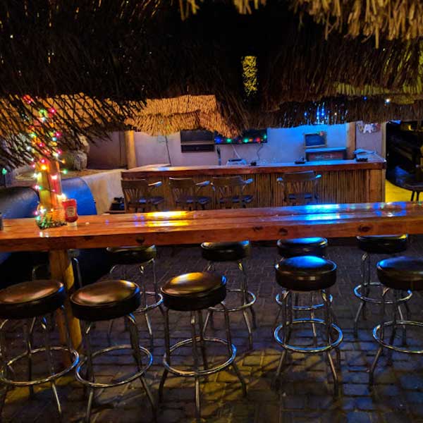 A row of empty bar stools lines a wooden counter in a dimly lit bar with string lights and festive decorations.