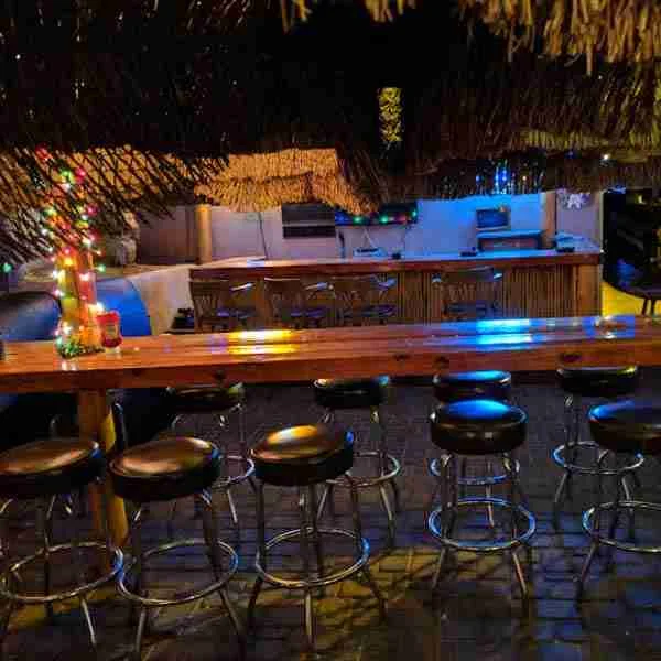 A row of empty bar stools lines a wooden counter in a dimly lit bar with string lights and festive decorations.