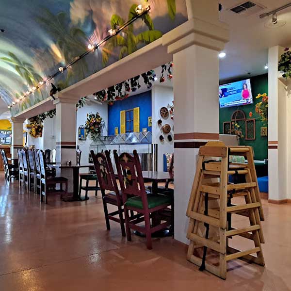 Restaurant interior with long tables and chairs, stacked wooden high chairs, colorful wall decor, ceiling mural, and a TV mounted on the wall.