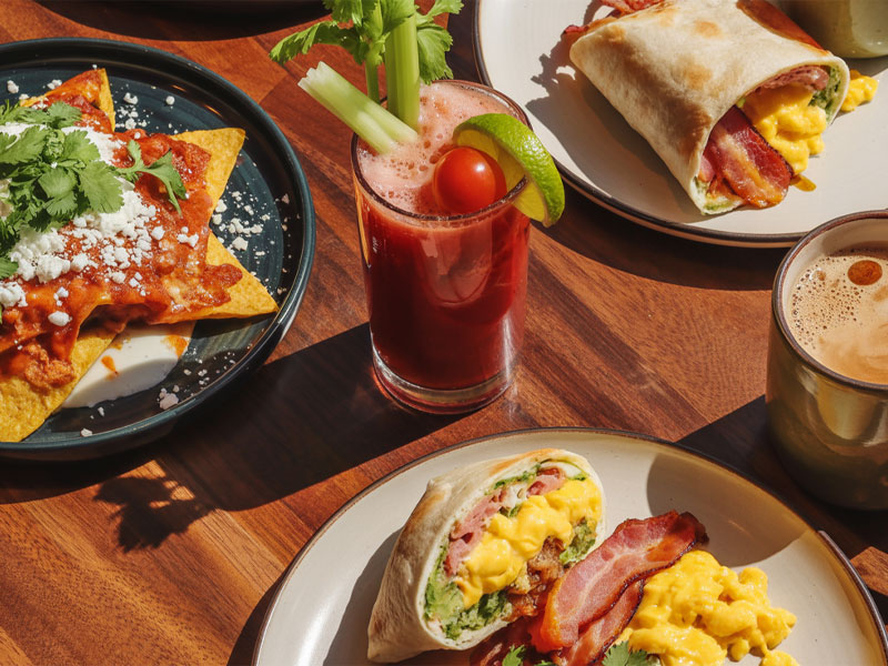 Plates with breakfast wraps, chilaquiles, a Bloody Mary garnished with celery and tomato, and a cup of coffee on a wooden table.