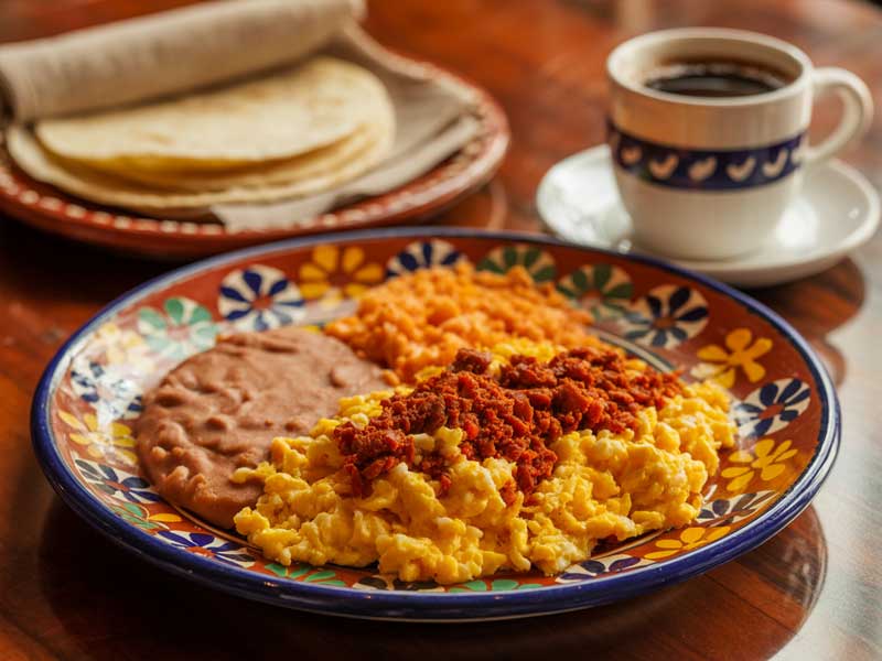 A plate with scrambled eggs topped with chorizo, refried beans, and Mexican rice, next to a cup of coffee, tortillas, and a folded newspaper.
