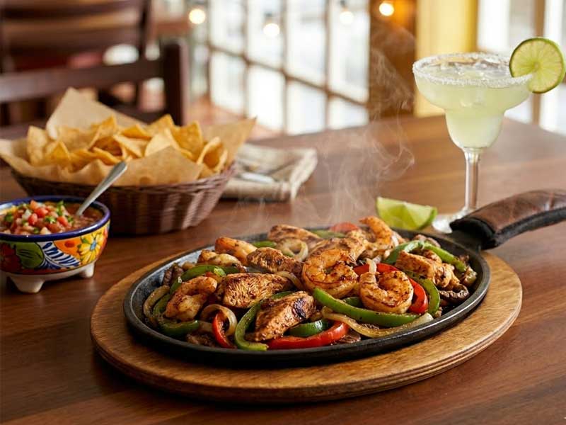 A sizzling platter of grilled shrimp and vegetables with lime, served with tortilla chips, salsa, and a margarita garnish with a lime slice on a wooden table.