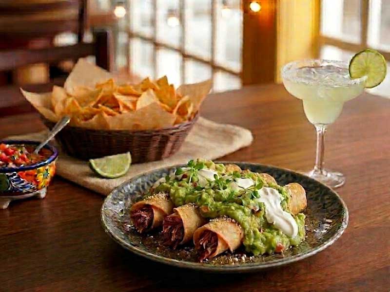 A plate of taquitos topped with guacamole and sour cream, a bowl of salsa, a basket of tortilla chips, and a margarita with a lime slice on a wooden table.