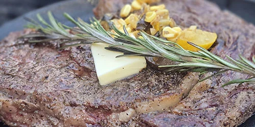 Grilled steak topped with a sprig of rosemary, a pat of butter, chopped vegetables, and cracked black pepper.