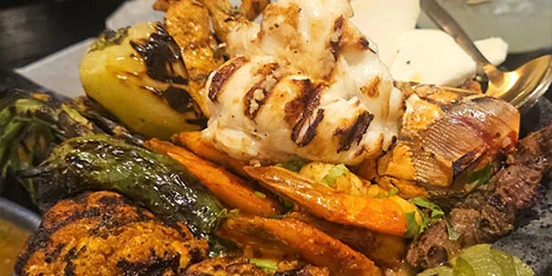 A close-up of assorted grilled meats and vegetables, including chicken, fish, peppers, carrots, and potatoes, served on a platter.