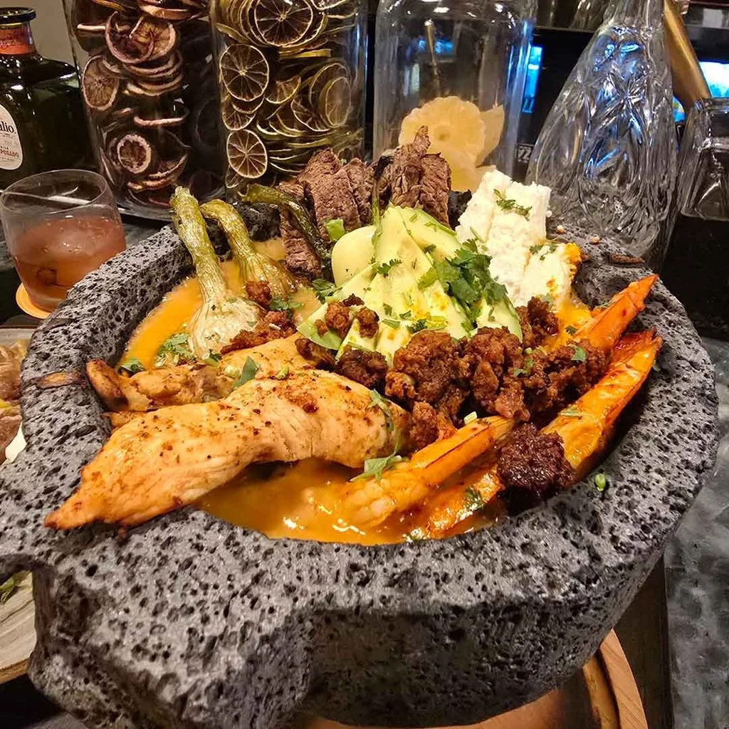 Molcajete bowl filled with grilled chicken, steak, chorizo, shrimp, avocado, cheese, and roasted vegetables in a sauce.